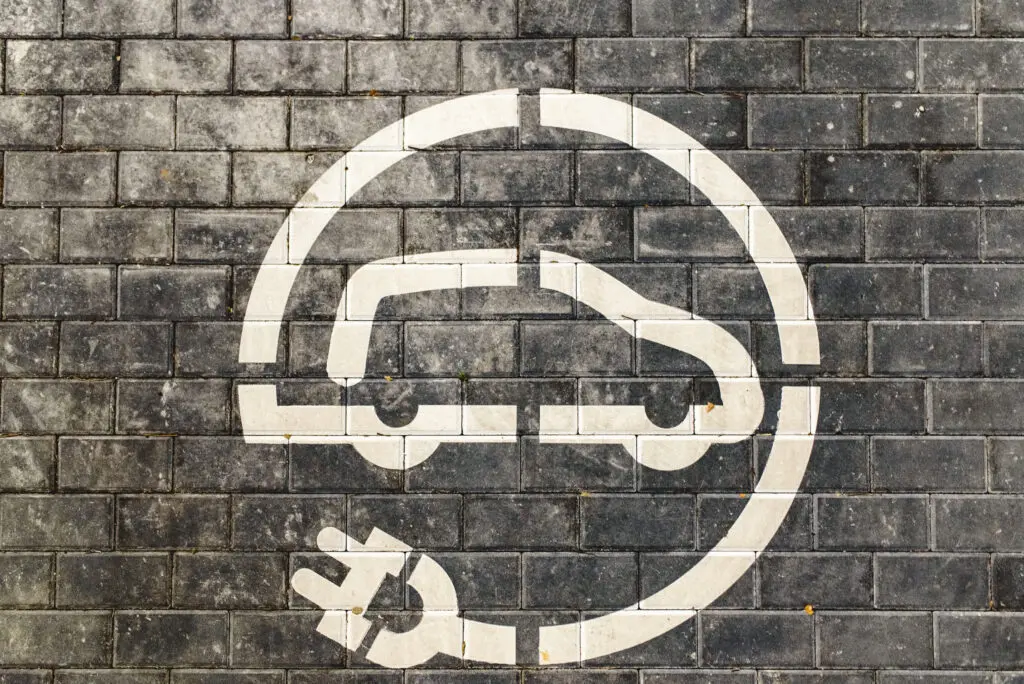 Graphic icon representing electric vehicle (EV) charging. The graphic displays an icon of a motor vehicle that has an electrical cord attached to the back of the vehicle and wrapping around the vehicle in a circular direction to complete the majority of a circle boundary for the graphic.